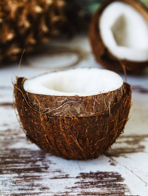 Eine grosse, braune Kokosnuss, welche halbiert ist und ein frisches weisses Fruchtfleisch zeigt. Im Hintergrund sieht man andere Kokosnüsse, welche verschwommen sind.